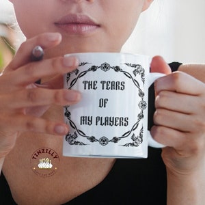 May include: White ceramic mug with the text "THE TEARS OF MY PLAYERS" in black, framed by a decorative border. The mug is held by a person wearing a black top.