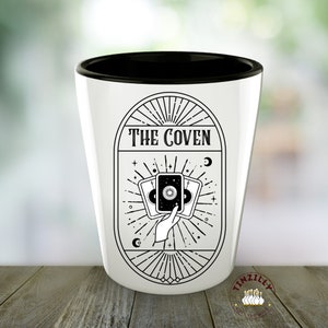 Op de afbeelding: Een wit keramiek shotglas met een zwarte rand. Het glas heeft een zwart-witte illustratie van een hand die drie tarotkaarten vasthoudt met de tekst "THE COVEN" boven de kaarten.