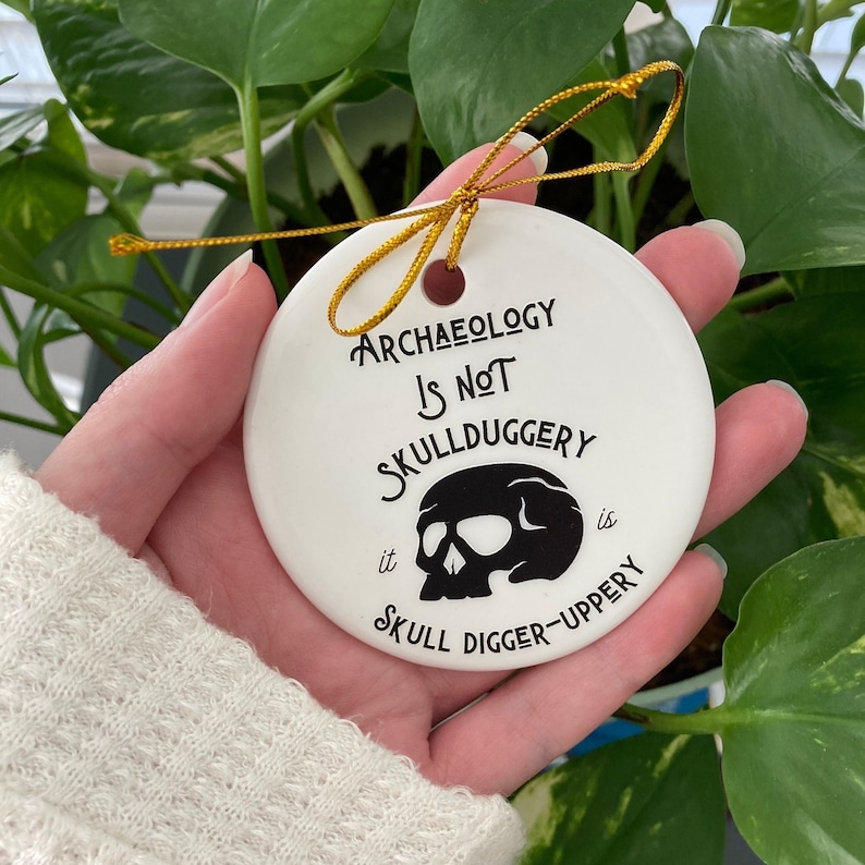May include: White ceramic ornament with black text and a skull graphic. The text reads "Archaeology is not skullduggery it is skull digger-uppery".