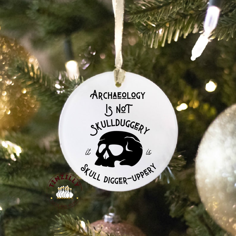 May include: White round Christmas ornament with black text that reads "Archaeology is not skullduggery it is skull digger-uppery" and a black skull graphic.