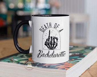 May include: White ceramic coffee mug with a black handle and black rim. The mug features a black graphic of a skeleton hand giving the middle finger with a ring on the finger. The text "Death of a Bachelorette" is printed on the mug.
