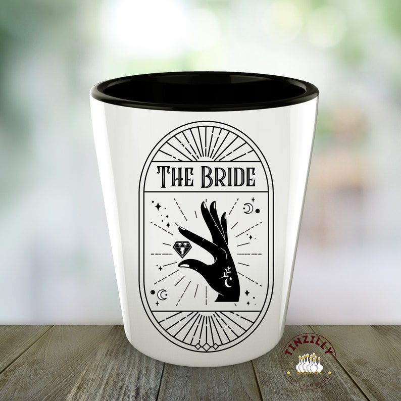 Op de afbeelding: Een wit keramiek shotglas met een zwarte rand. Het glas heeft een zwart-witte afbeelding van een hand die een diamant vasthoudt met de tekst "THE BRIDE" erboven.