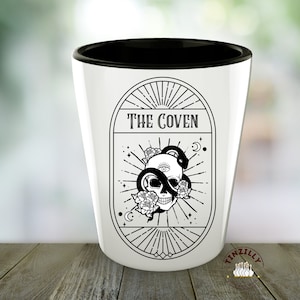 Op de afbeelding: Een wit keramiek shotglas met een zwarte rand. Het glas heeft een zwart-witte illustratie van een schedel met een slang eromheen gewikkeld. De illustratie is omgeven door een cirkel met de tekst "THE COVEN" bovenaan.
