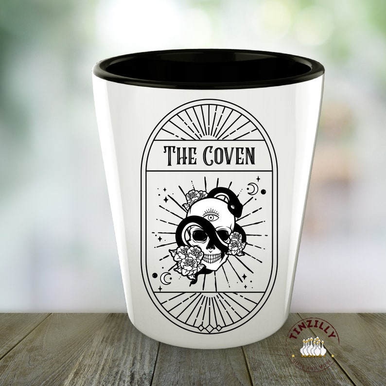 Op de afbeelding: Een wit keramiek shotglas met een zwarte rand. Het glas heeft een zwart-witte illustratie van een schedel met een slang eromheen gewikkeld. De illustratie is omgeven door een cirkel met de tekst "THE COVEN" bovenaan.