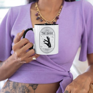 May include: White ceramic mug with a black rim and handle. The mug has a black and white graphic of a hand holding a diamond ring with the text "THE BRIDE" above it.