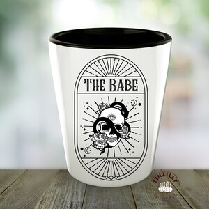 Op de afbeelding: Een wit keramiek shotglas met een zwarte rand. Het glas heeft een zwart-wit grafisch ontwerp van een schedel met een slang eromheen, omringd door bloemen en sterren. De tekst "THE BABE" is boven de schedel gedrukt.