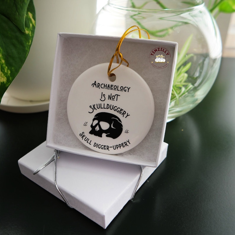 May include: White ceramic ornament with a black skull and the text "Archaeology is not skullduggery it is skull digger-uppery".