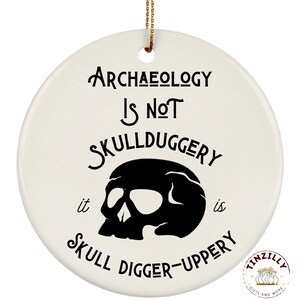 May include: White ceramic ornament with black text that reads "Archaeology is not skullduggery it is skull digger-uppery". A black skull graphic is in the center of the ornament.