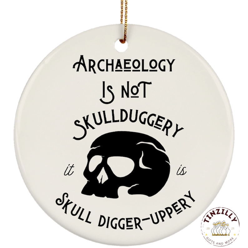 May include: White ceramic ornament with black text that reads "Archaeology is not skullduggery it is skull digger-uppery". A black skull graphic is in the center of the ornament.