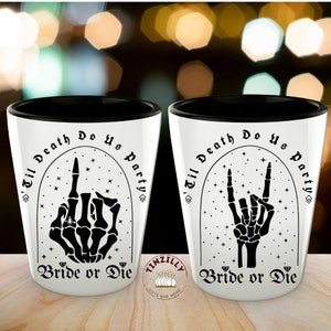 May include: Two white shot glasses with black rims. The glasses feature a black and white graphic of a skeleton hand with the text "Til Death Do Us Party" and "Bride or Die".