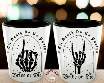 May include: Two white shot glasses with black rims. The glasses have a black and white graphic of a skeleton hand with the text "Til Death Do Us Party" and "Bride or Die".
