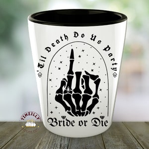 May include: White ceramic shot glass with a black rim and a black graphic of a skeleton hand giving the middle finger with a diamond ring on the ring finger. The text "'Til Death Do Us Party" is written in a black gothic font above the hand and the text "Bride or Die" is written below the hand.
