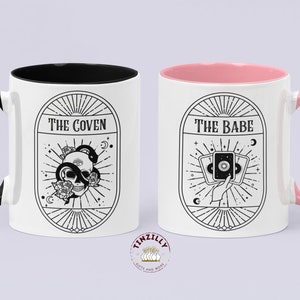May include: Two white ceramic mugs with black and pink accents. The mug on the left has a black handle and a black rim. The mug on the right has a pink handle and a pink rim. Both mugs feature black and white illustrations with text. The mug on the left says "The Coven" and features a skull with a snake wrapped around it. The mug on the right says "The Babe" and features a hand holding three tarot cards.