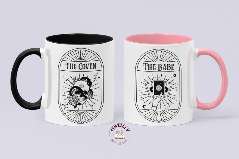 May include: Two white ceramic mugs with black and pink accents. The mug on the left has a black handle and a black rim. The mug on the right has a pink handle and a pink rim. Both mugs feature black and white illustrations with text. The mug on the left says "The Coven" and features a skull with a snake wrapped around it. The mug on the right says "The Babe" and features a hand holding three tarot cards.