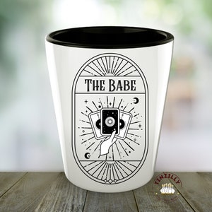 Op de afbeelding: Een wit keramiek shotglas met een zwarte rand. Het glas heeft een zwart-witte illustratie van een hand die drie tarotkaarten vasthoudt met de tekst "THE BABE" boven de kaarten.