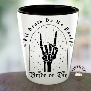 May include: White ceramic shot glass with a black rim and a black design. The design features a skeleton hand making the rock and roll hand gesture with the words "'Til Death Do Us Party" and "Bride or Die" in a gothic font.