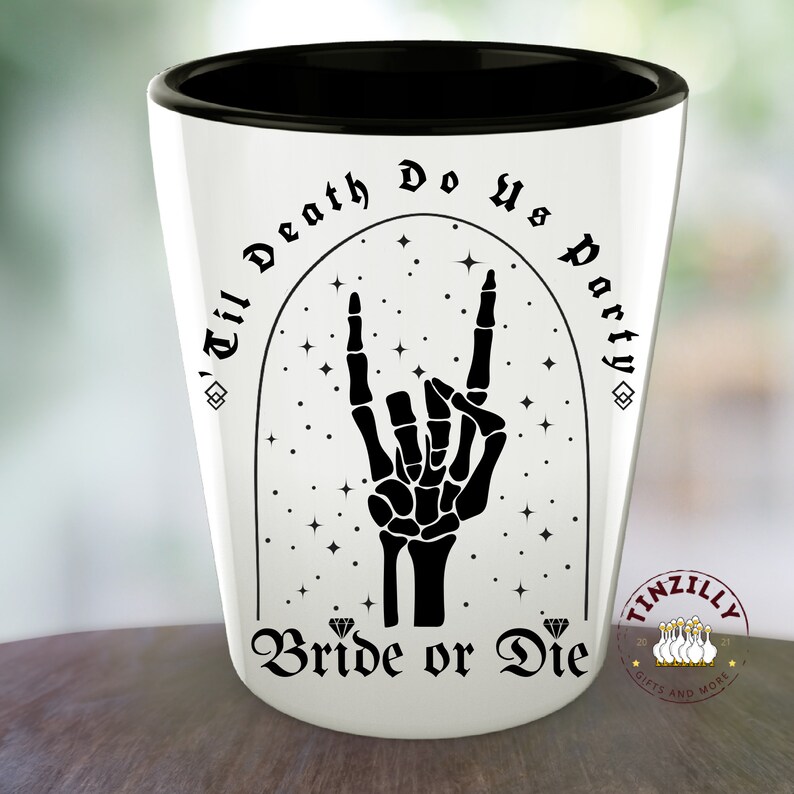 May include: White ceramic shot glass with a black rim and a black design. The design features a skeleton hand making the rock and roll hand gesture with the words "'Til Death Do Us Party" and "Bride or Die" in a gothic font.