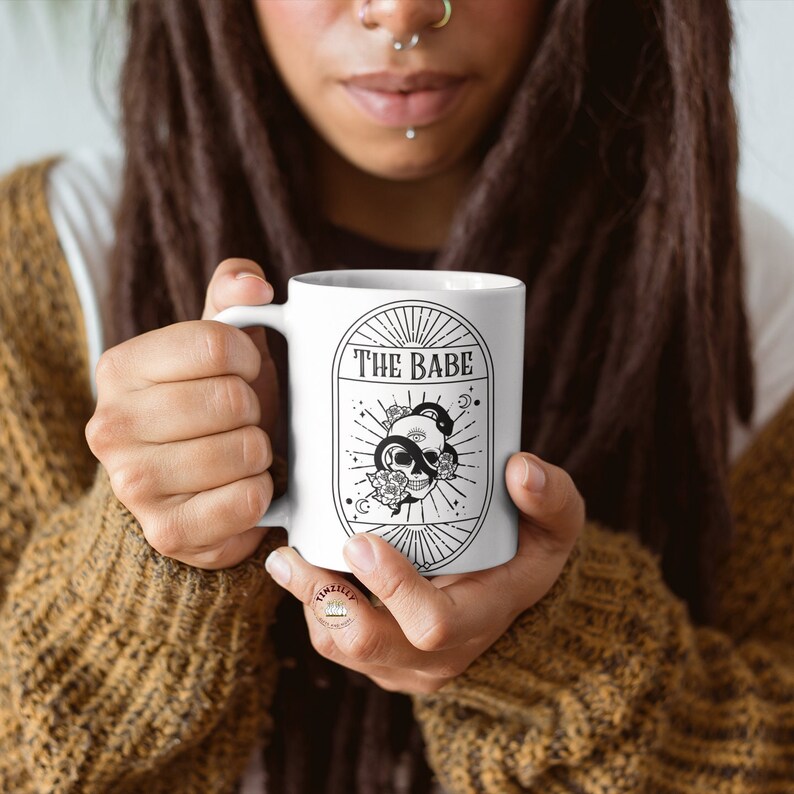 May include: White ceramic mug with a black and white illustration of a skull with a snake wrapped around it. The text "THE BABE" is printed on the mug.