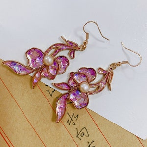 Puede incluir: Un par de pendientes de oro con diseños de flores iridiscentes rosas y detalles de perlas.