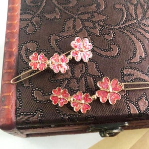 Puede incluir: Dos pinzas para el pelo doradas con detalles de flores rosas. Cada pinza tiene tres flores conectadas por pequeñas bolas dorées. Las flores tienen un acabado brillante e iridiscente.