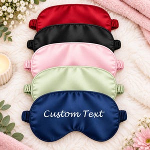 May include: A stack of five satin sleep masks in red, black, light pink, light green, and navy blue. Each mask features a gathered elastic strap. The navy blue mask at the bottom has the words Custom Text printed in white script lettering.