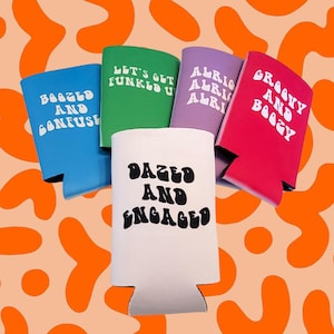 May include: A collection of colorful beverage can coolers. The coolers are blue, green, purple, red, and white. Each cooler has a different phrase in white, including "Dazed and Engaged" and "Groovy and Boogy". The background is orange.