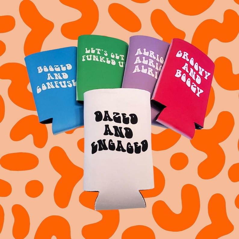 May include: A collection of colorful beverage can coolers. The coolers are blue, green, purple, red, and white. Each cooler has a different phrase in white, including "Dazed and Engaged" and "Groovy and Boogy". The background is orange.