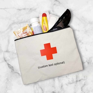 May include: A white canvas zippered pouch with a red cross and the text "(custom text optional)". The pouch is filled with various items, including a yellow tube of ointment, a clear bottle of pills, and a packet of vitamin C.