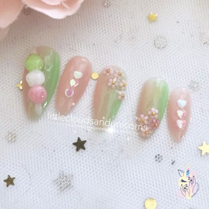 Puede incluir: Conjunto de seis uñas postizas de color verde pastel y rosa con diferentes diseños. Una uña tiene un diseño dango rosa y blanco, otra tiene corazones rosa y blanco, otra tiene flores rosas y otra tiene flores rosas con detalles dorados. Las uñas están sobre un fondo blanco con estrellas doradas.