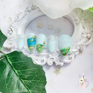 Könnte beinhalten: Fünf hellblaue, mandelförmige Kunstnägel mit verschiedenen Designs. Ein Nagel hat ein kleines silbernes und weißes Design mit einer Perle. Ein Nagel hat ein kleines silbernes und weißes Design mit einer Perle. Ein Nagel hat ein kleines silbernes und weißes Design mit einer Perle. Ein Nagel hat ein kleines silbernes und weißes Design mit einer Perle. Ein Nagel hat ein kleines silbernes und weißes Design mit einer Perle. Die Nägel werden auf einem weißen Hintergrund mit einem weißen Rahmen präsentiert.