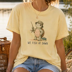 May include: A pale yellow short-sleeve t-shirt featuring a graphic of a turtle wearing a bucket hat, holding a fishing rod, and standing next to a tackle box. Below the illustration, the text reads WE FISH AT DAWN in black capital letters.