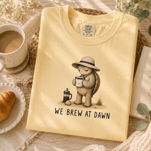 May include: A pale yellow t-shirt featuring a graphic of a turtle wearing a sun hat, holding a steaming mug of coffee next to a French press. The text "WE BREW AT DAWN" is printed below the illustration. The shirt has a Comfort Colors brand label.
