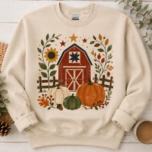 May include: A cream-colored crewneck sweatshirt featuring a graphic print of a red barn, a wooden fence, and three pumpkins in orange, white, and green. The design is surrounded by autumn leaves, sunflowers, and stars, with a Gildan brand label.