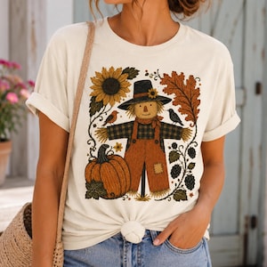 May include: A cream-colored short-sleeve t-shirt featuring a graphic print of a scarecrow wearing a hat, standing next to a large orange pumpkin, a sunflower, autumn leaves, and small birds, with a woven bag strap visible on the shoulder.