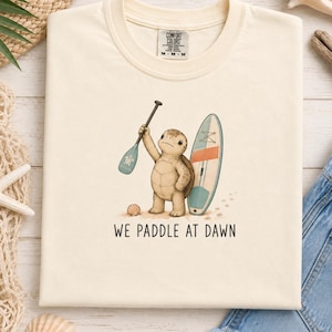 May include: A cream-colored t-shirt featuring a graphic of a turtle standing on sand, holding a paddle, and standing next to a surfboard. The text "WE PADDLE AT DAWN" is printed below the illustration. The shirt has a Comfort Colors brand label.