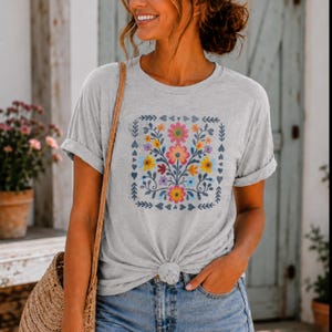 May include: A light gray short-sleeved t-shirt featuring a square graphic print of colorful flowers in shades of pink, yellow, orange, and blue, accented with dark blue leaves and small heart shapes arranged in a symmetrical botanical pattern.