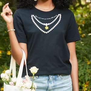 May include: Black t-shirt with a pearl necklace graphic and the text "You can't ever go wrong with pearls." The necklace design includes white pearls and a yellow flower pendant. The shirt is paired with blue jeans.