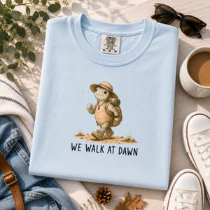 May include: A light blue t-shirt featuring a graphic of a cartoon turtle wearing a tan sun hat and a backpack, walking on a dirt path. Below the turtle, the text reads WE WALK AT DAWN in black capital letters. The shirt has a Comfort Colors label.