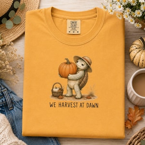 May include: A mustard yellow t-shirt featuring a graphic of a turtle wearing a straw hat and holding a large orange pumpkin. Next to the turtle is a small woven basket containing a pumpkin. The text WE HARVEST AT DAWN is printed below.