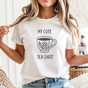 May include: White t-shirt with the text "MY CUTE TEA SHIRT" above a graphic of a teacup with a floral design and a tea bag. The shirt is a short-sleeved crew neck.