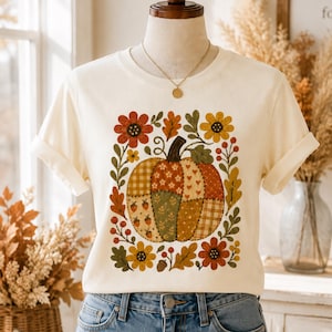May include: A cream-colored short-sleeve t-shirt featuring a central graphic of a patchwork-style pumpkin surrounded by autumn-themed illustrations including red and yellow flowers, green leaves, berries, and a small brown acorn.