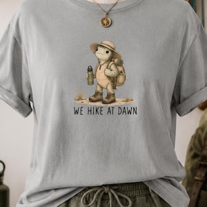 May include: A light gray t-shirt featuring a graphic of a turtle wearing a sun hat, a backpack, and hiking boots while holding a water bottle. Below the turtle, the text reads WE HIKE AT DAWN in black capital letters. The shirt has short sleeves.