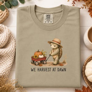 May include: A light olive green t-shirt featuring a graphic of a turtle wearing a straw hat and pulling a red wagon filled with pumpkins and a sunflower. The text below the graphic reads WE HARVEST AT DAWN. The shirt has a Comfort Colors label.