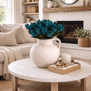 May include: A living room with a round coffee table holding a large, textured vase of teal tulips. A candle and tray are also on the table. The room has a neutral colour scheme, a fireplace, and shelves, creating a cosy atmosphere.