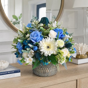 May include: A floral arrangement featuring blue and white roses, a white gerbera daisy, and dark blue thistle accents. The flowers are arranged in a round, textured grey stone-like pot with vertical grooves, creating a dense, rounded display.
