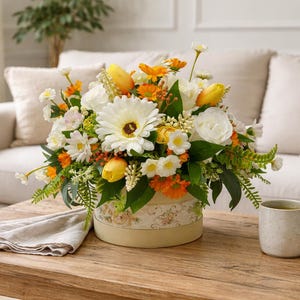 Op de afbeelding: Een bloemstuk in een ronde, crèmekleurige doos met een bloemenmotief, met witte gerbera's, gele tulpen en oranje bloemen. Een beige mok en een gevouwen linnen servet liggen op een houten tafel.