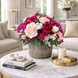 May include: A colourful floral display with roses and other flowers in pinks, reds, and white, in a textured vase. The arrangement is on a marble-topped coffee table with a gold base, with books and a decorative object.
