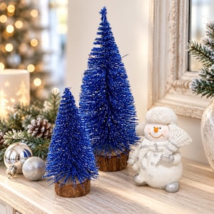 May include: Two conical bottle brush trees in a vibrant royal blue colour with glittery texture, each mounted on a natural wood slice base, positioned next to a small white snowman figurine wearing a hat and scarf while holding a broom.