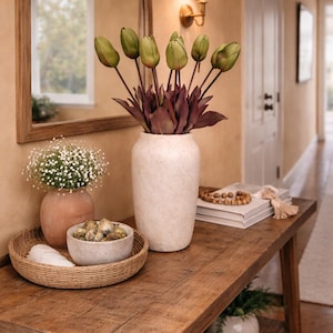May include: A wooden console table displaying home decor. A tall white vase contains green tulips and purple leaves. Other items include a small vase with white flowers, a woven tray, and a decorative bowl. The table is against a beige wall.