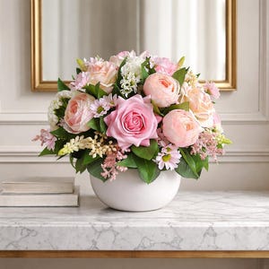 Peut inclure: Un arrangement floral composé de roses roses et blanches, de marguerites et d'autres fleurs dans un vase rond en céramique blanche. L'ensemble est posé sur une surface à motif marbré, avec un miroir encadré d'or en arrière-plan.
